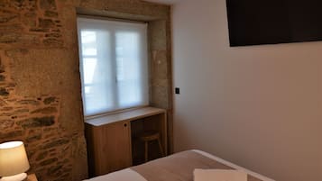 Un dormitorio con pared de piedra, un escritorio de madera, una ventana con persianas, una cama con manta blanca y un televisor fijado a la pared.