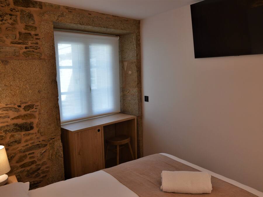 Un dormitorio con pared de piedra, un escritorio de madera, una ventana con persianas, una cama con manta blanca y un televisor fijado a la pared.