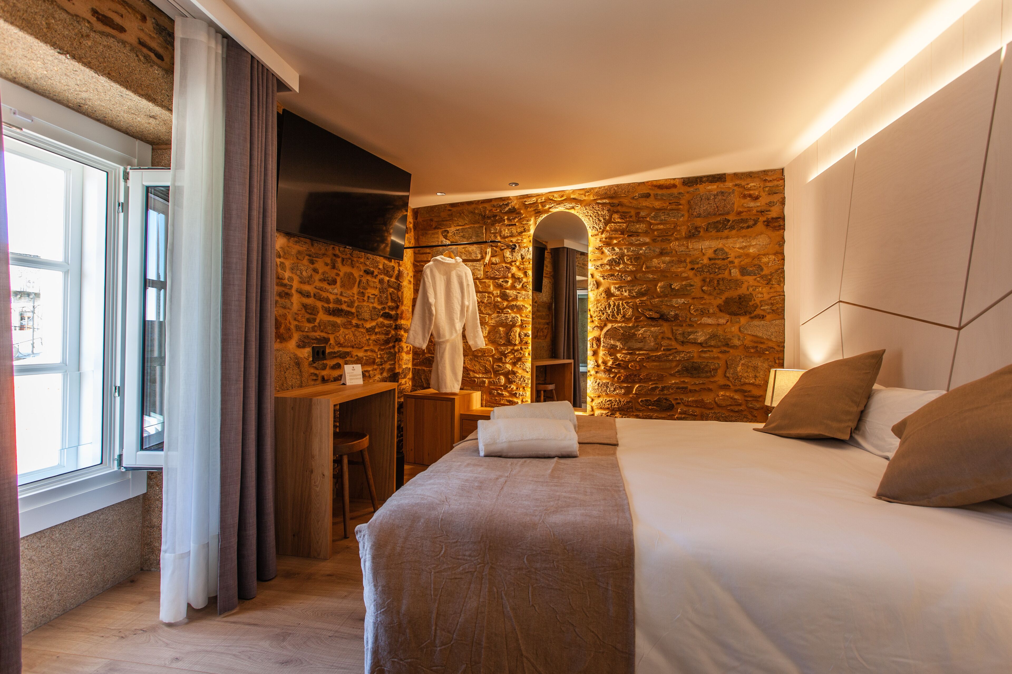 Ein Hotelzimmer mit Steinwand, einem Bett mit Kissen, einem Schreibtisch mit Stuhl und einem auf einem Haken hängenden Bademantel.
