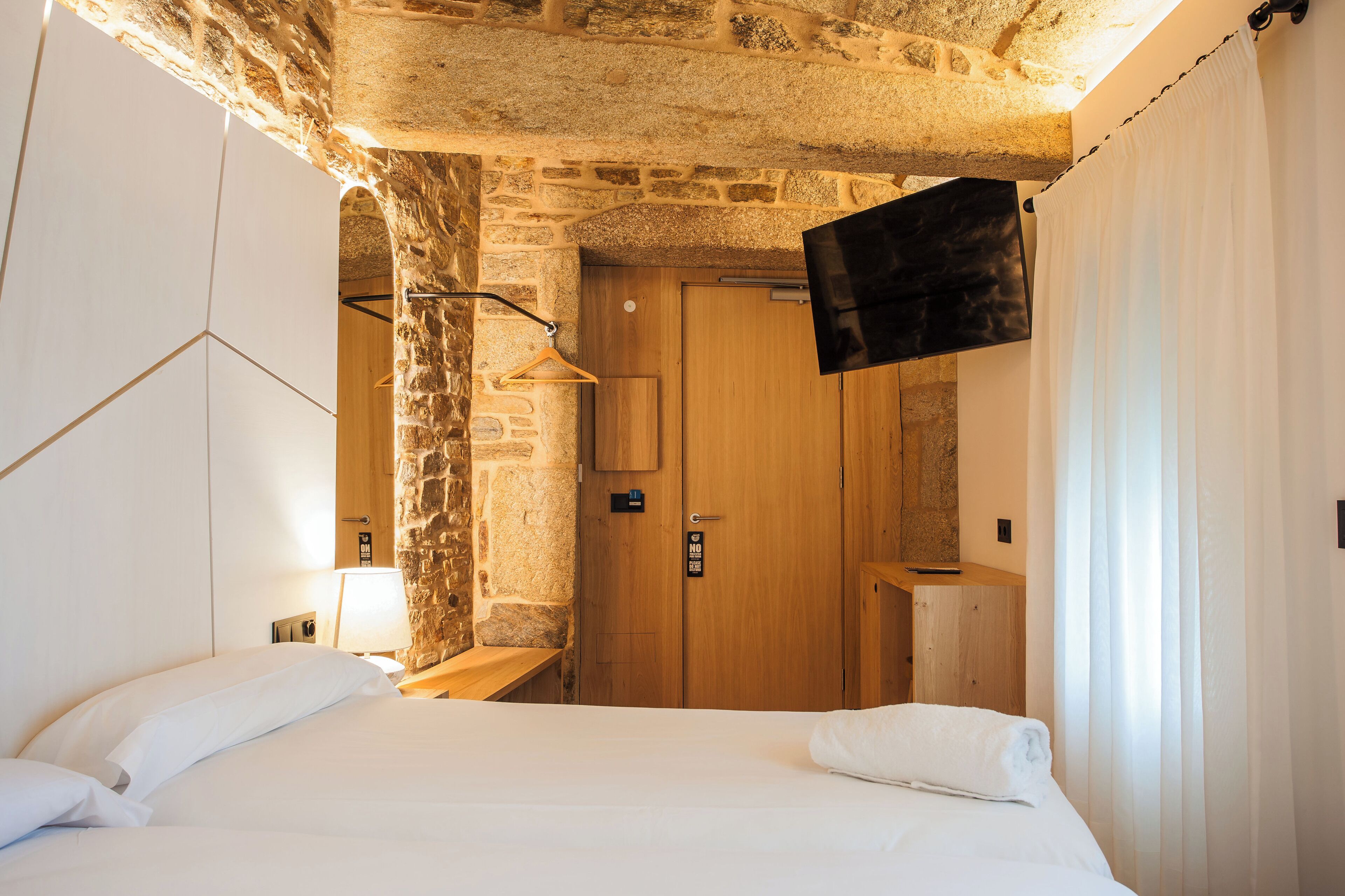 A hotel room with a stone wall, a wooden door, a flat-screen TV, a bed with white linens, and a wooden nightstand.
