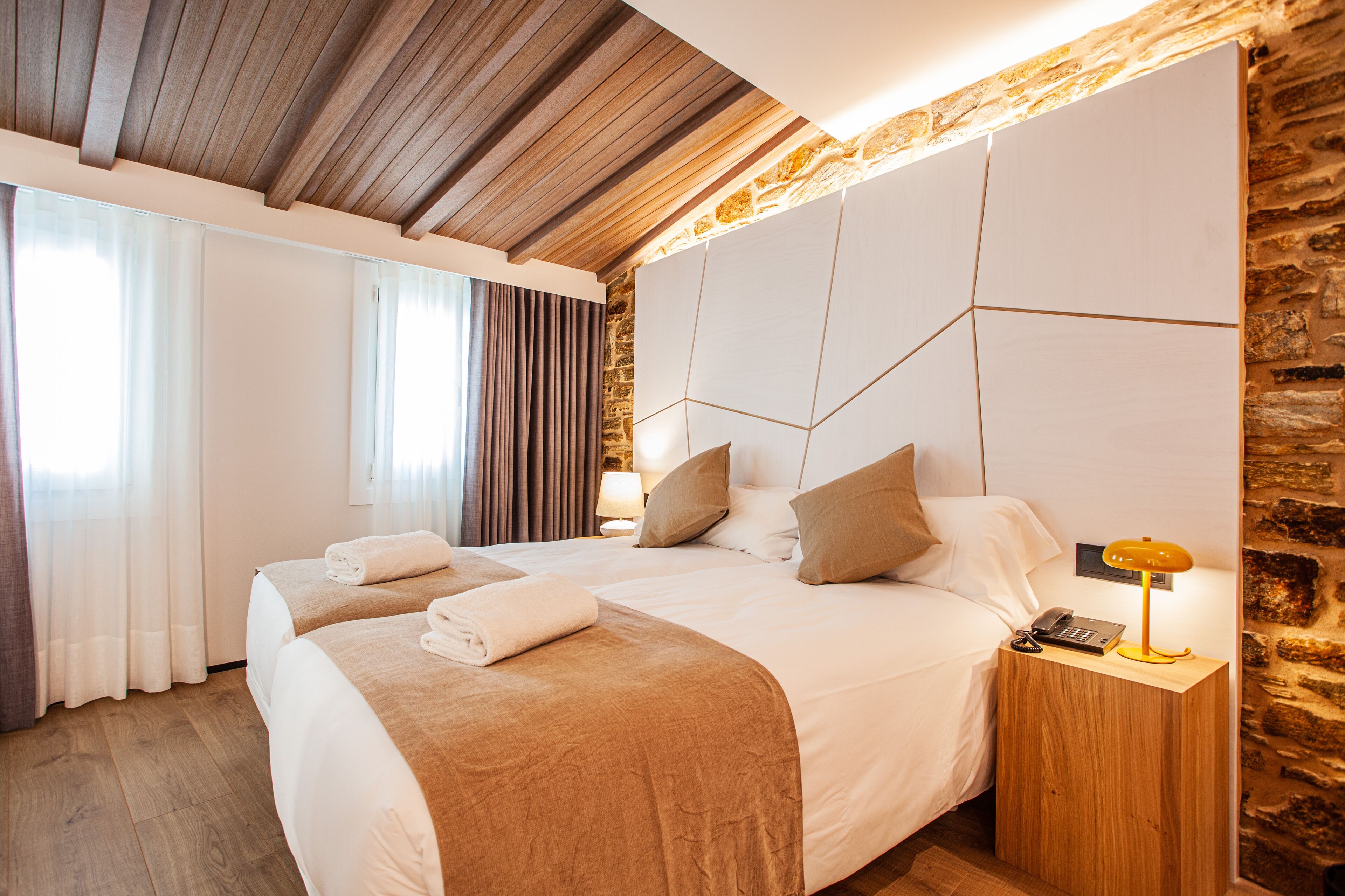 Habitación de hotel con dos camas, techo de madera, pared de piedra y una mesita de noche con lámpara.