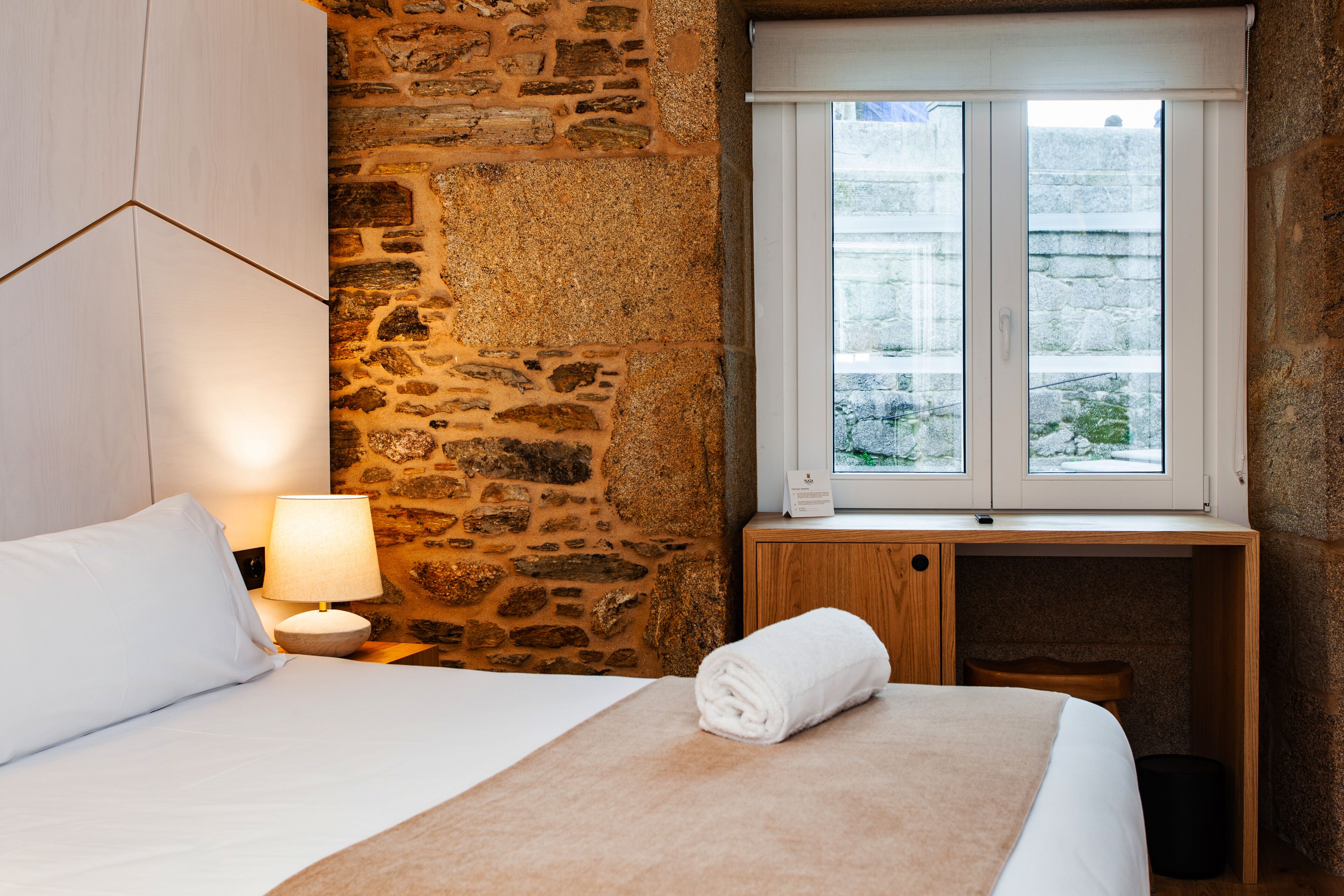 Une chambre d’hôtel avec un mur en pierre, un lit avec une literie blanche, un bureau en bois et une fenêtre donnant sur l’extérieur.