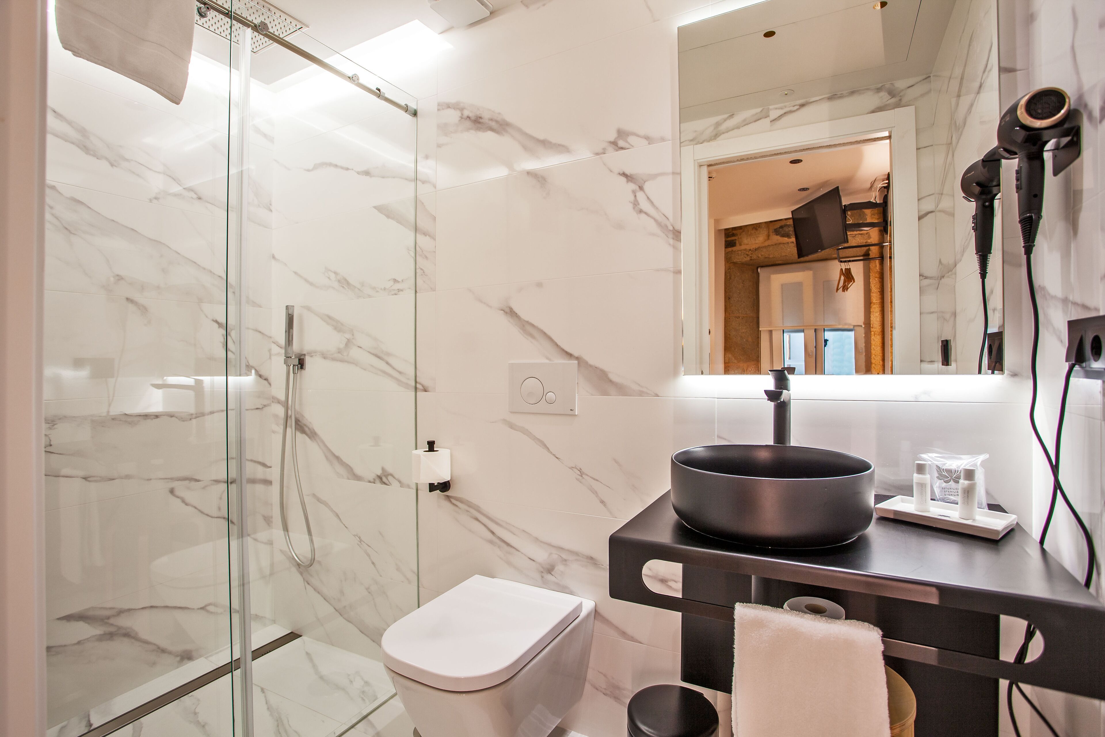 A modern bathroom with a glass-enclosed shower, a marble-tiled wall, a black sink with a bowl, and a mirror with a hairdryer.