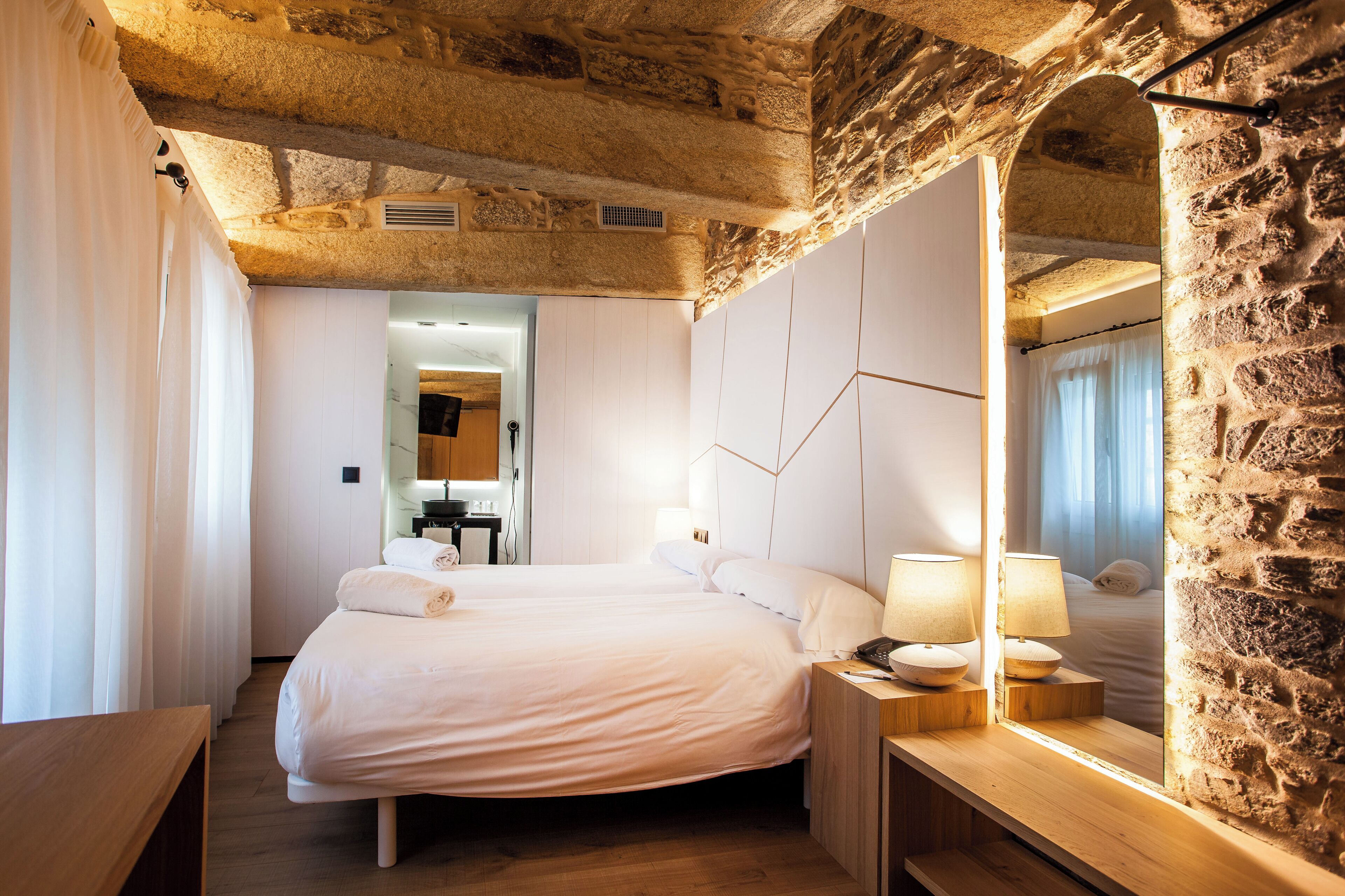 A bedroom with a stone wall, a wooden bed frame, and a nightstand with lamps.