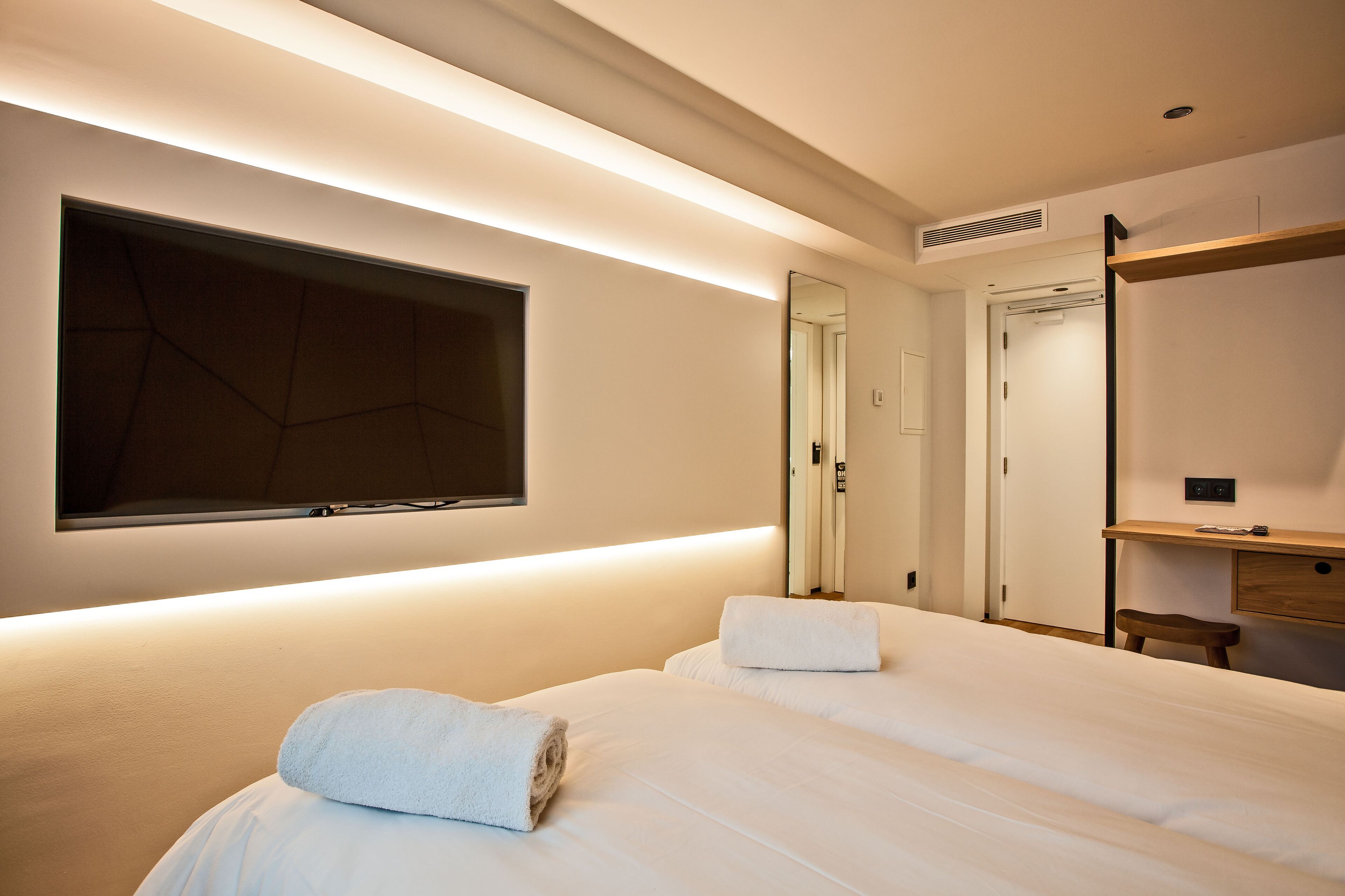 A modern hotel room with two beds, a large mirror, a TV mounted on the wall, and a wooden desk.