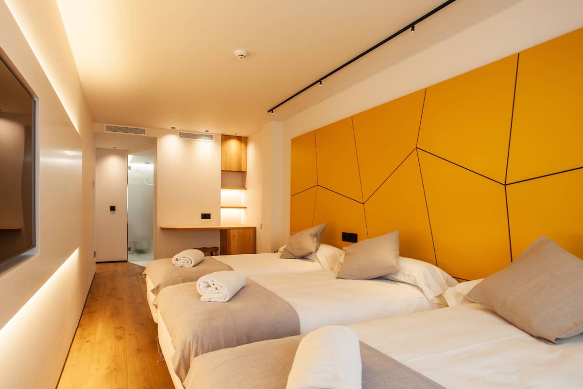 A modern hotel room with two beds, a wooden floor, a yellow accent wall, and a small desk area.