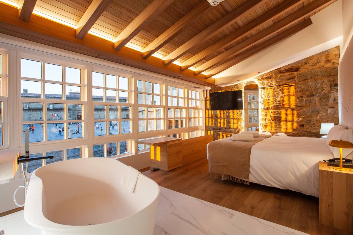 A bedroom with a large window, a bathtub, a bed, and a stone wall.