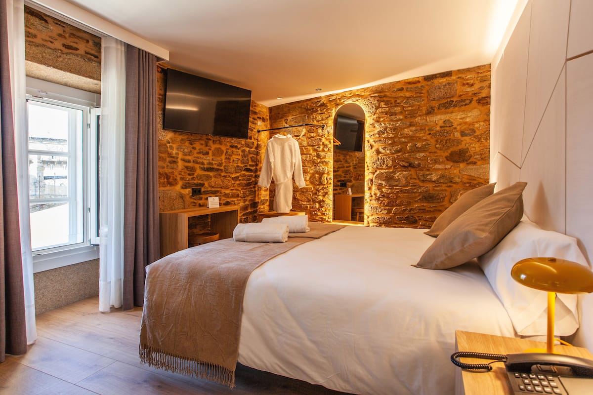 A hotel room with a stone wall, a bed with white linens, a bedside table with a lamp, and a robe hanging on a rack.
