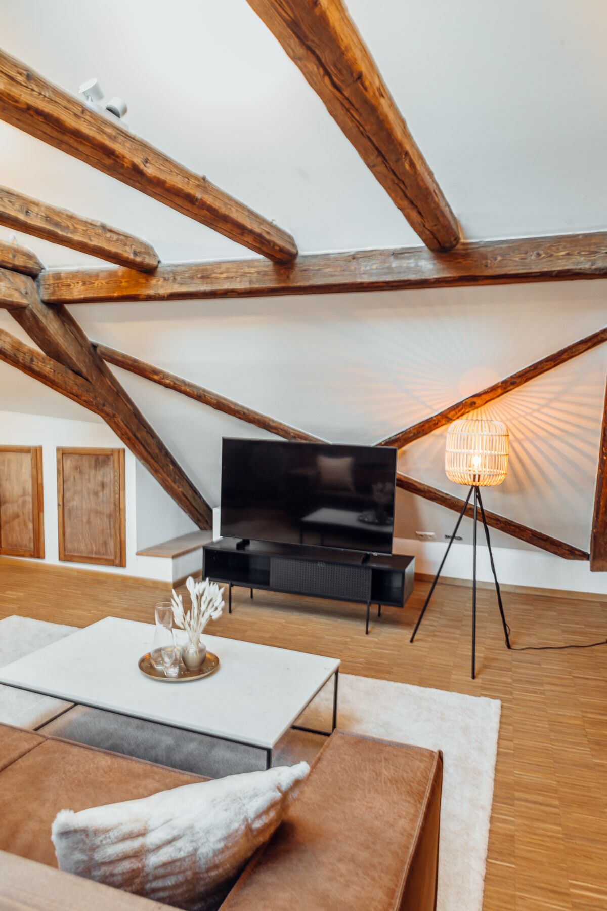 A modern loft bedroom with exposed wooden beams, a flat-screen TV, a standing lamp, and a minimalist decor.