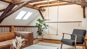 A loft-style room with exposed wooden beams, a hanging swing, a brown leather couch, a grey armchair, and a large potted plant.