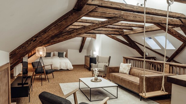 A spacious loft bedroom with a high ceiling, exposed wooden beams, a bed, a desk, a sofa, and a swing chair.