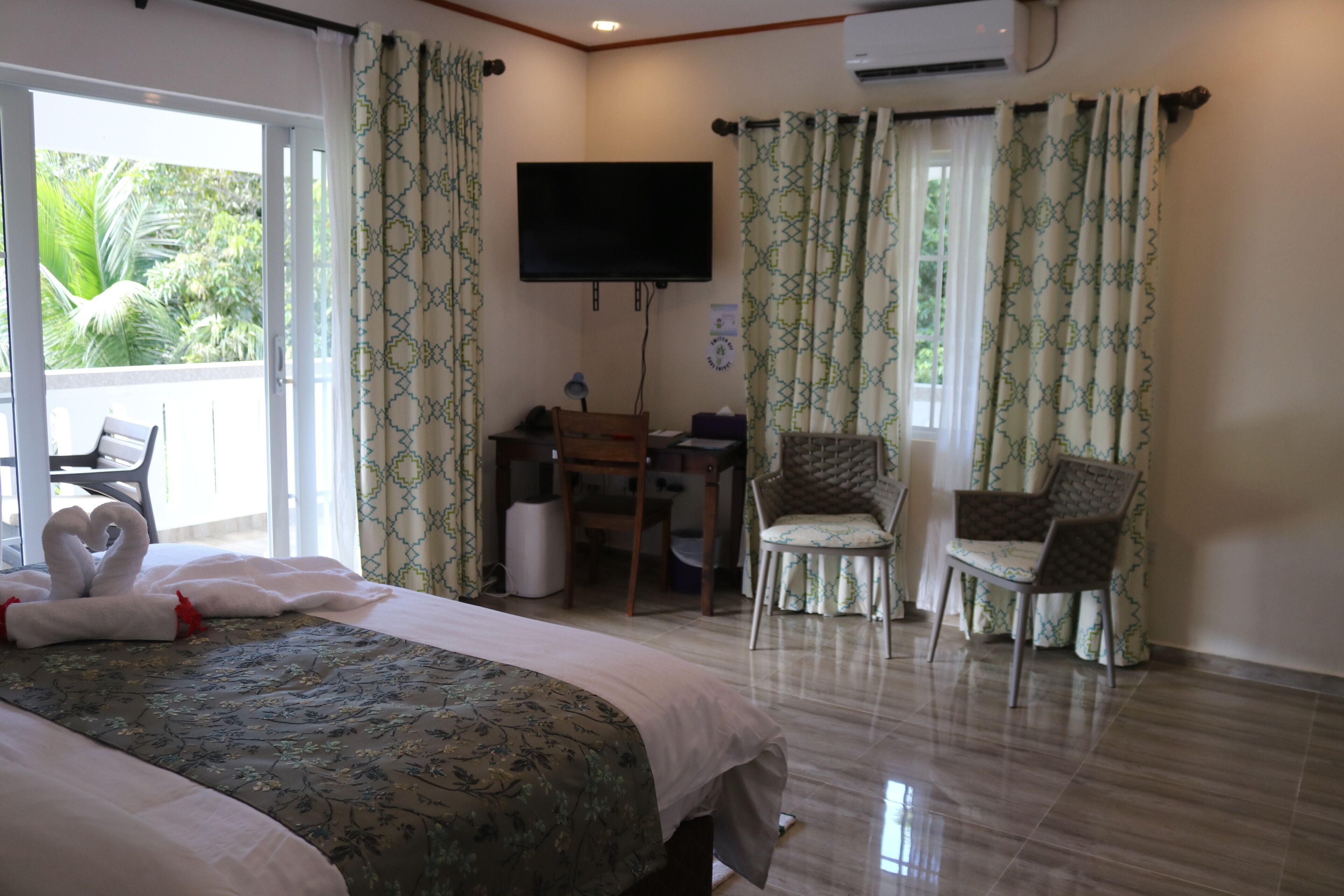 Habitación de hotel con cama, escritorio, dos sillas, televisor y balcón con cortinas.