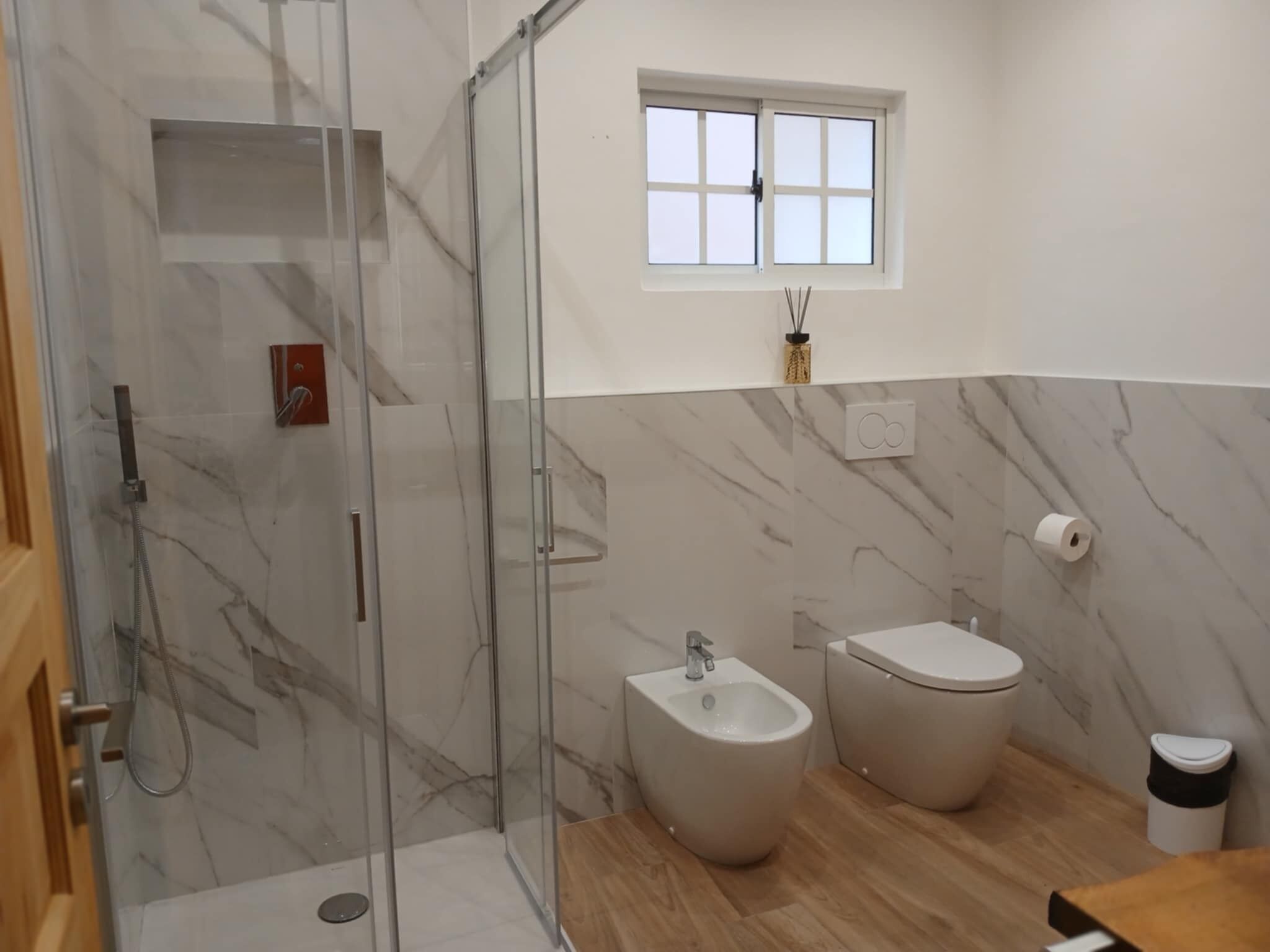 A modern bathroom with a glass-enclosed shower, a bidet, a toilet, and a window.