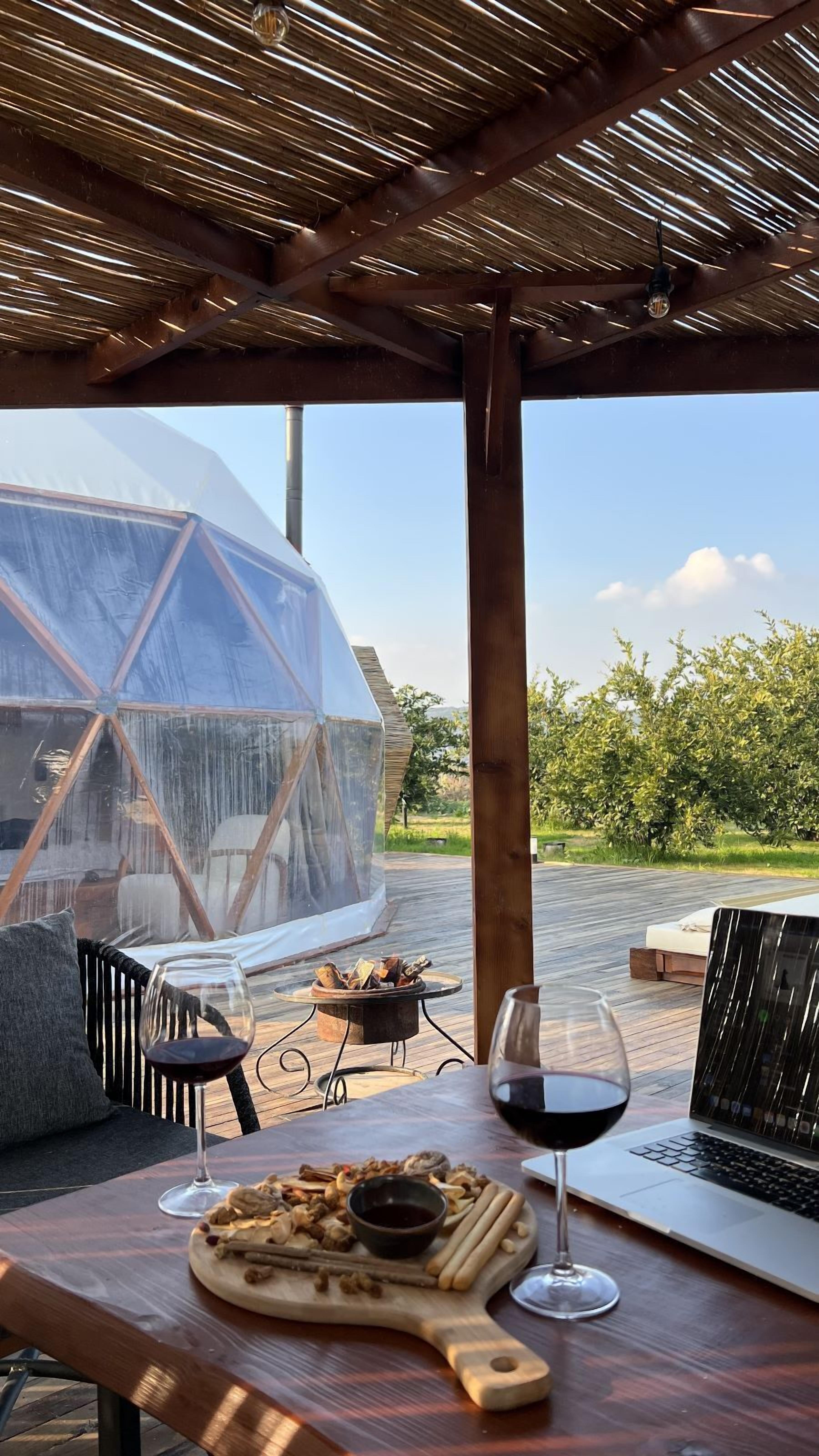 Un tavolo di legno con un laptop, bicchieri da vino e un tagliere con cibo. Sullo sfondo, una zona riparata con una cupola trasparente e vista sul verde.