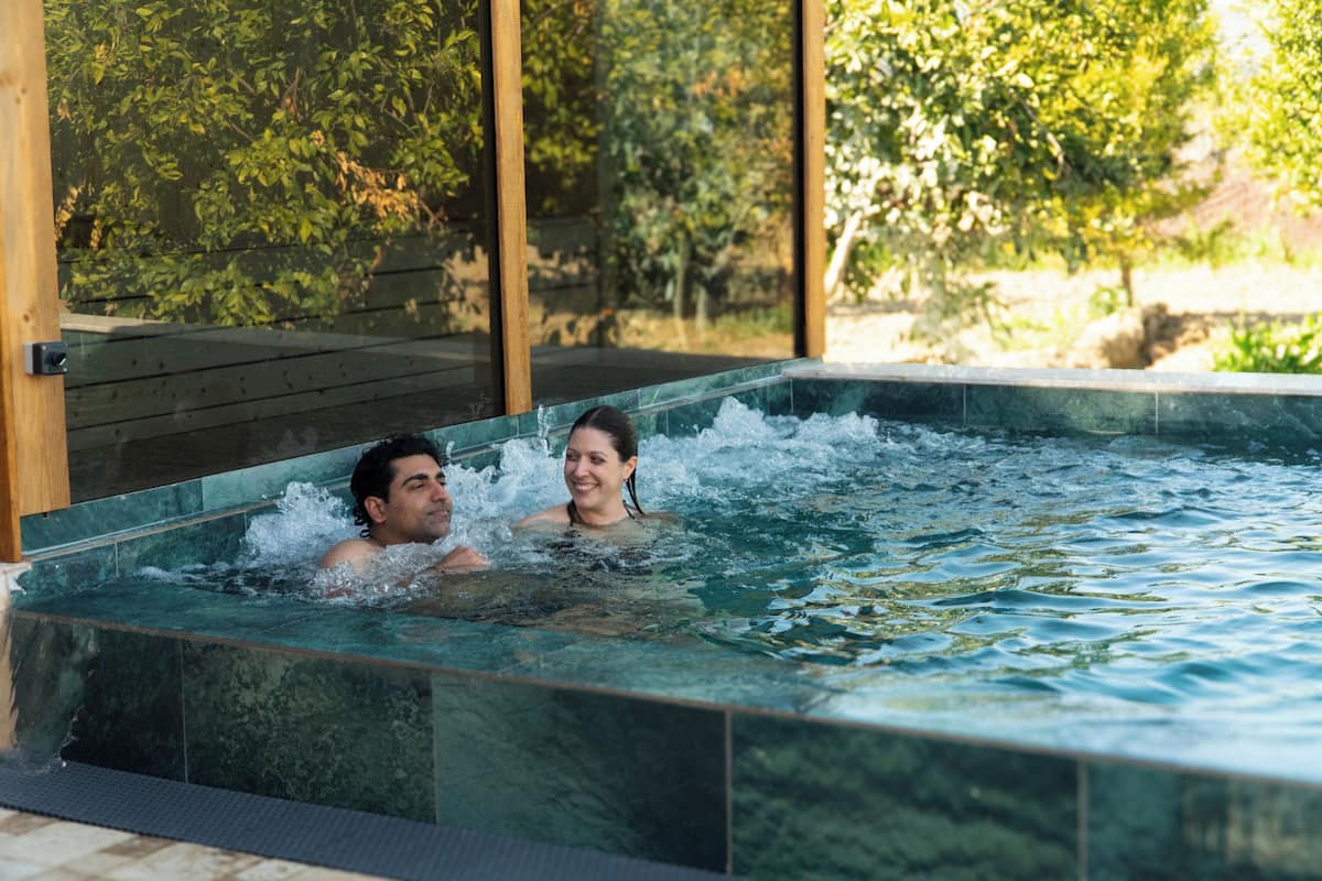 Un couple se détend dans un jacuzzi rectangulaire, entouré d’une terrasse en bois et offrant une vue sur la verdure.
