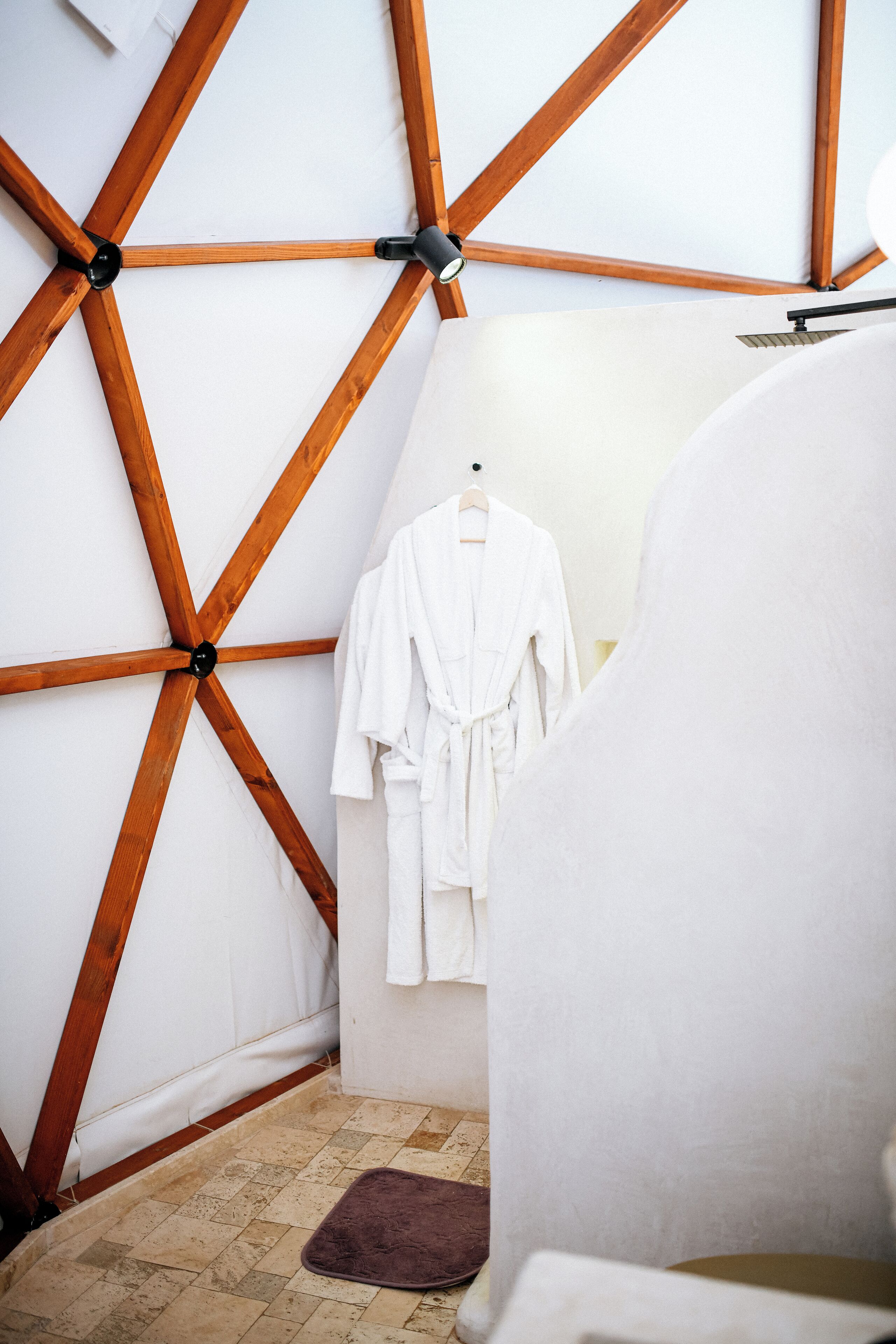 A room with a wooden ceiling structure, a white robe hanging on a hook, and a brown rug on the floor.