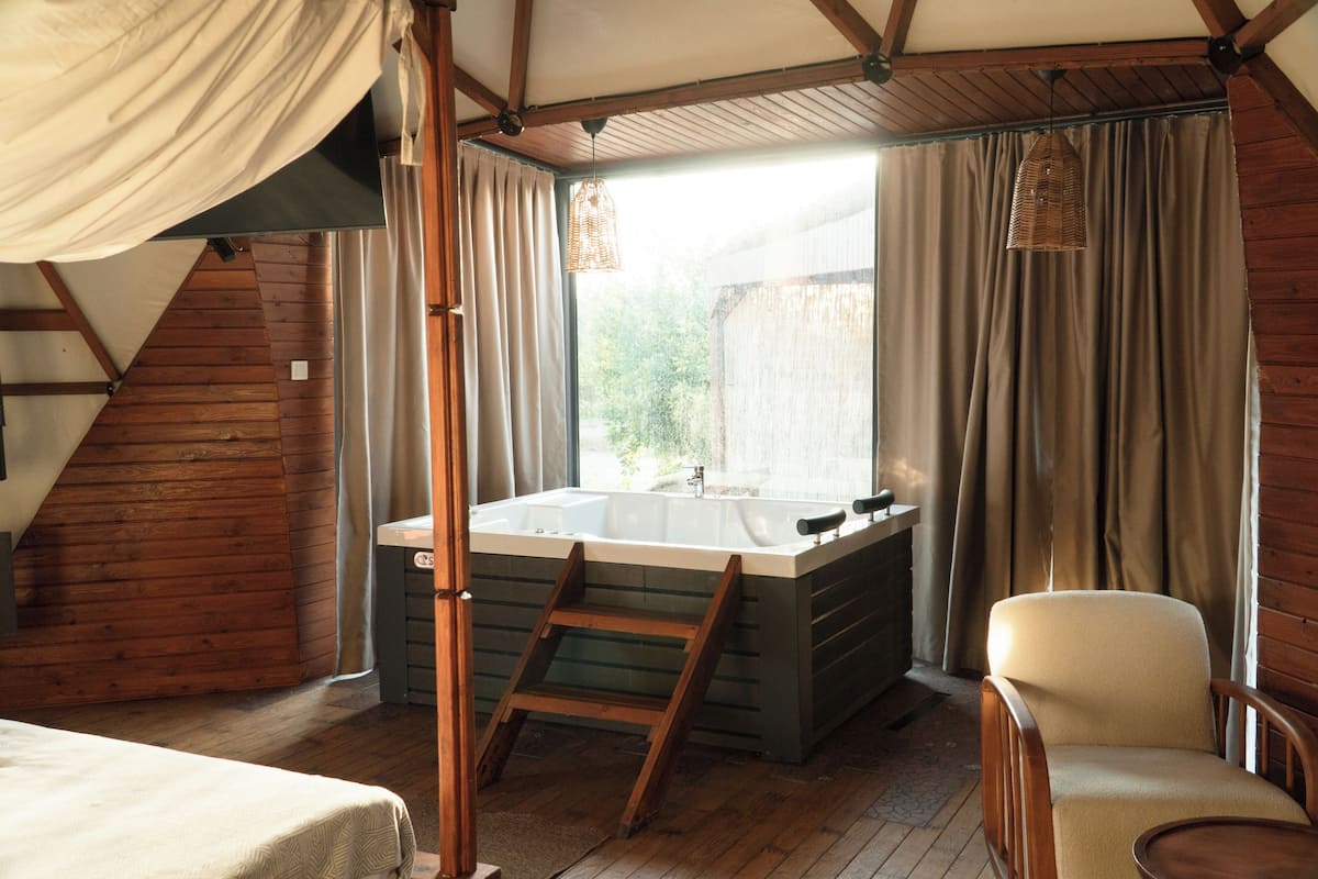 Une pièce avec un plancher en bois, une grande fenêtre avec des rideaux, une baignoire, une chaise et une échelle.