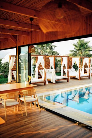 A modern pool area with wooden deck, lounge chairs, and a glass-enclosed space with curtains.