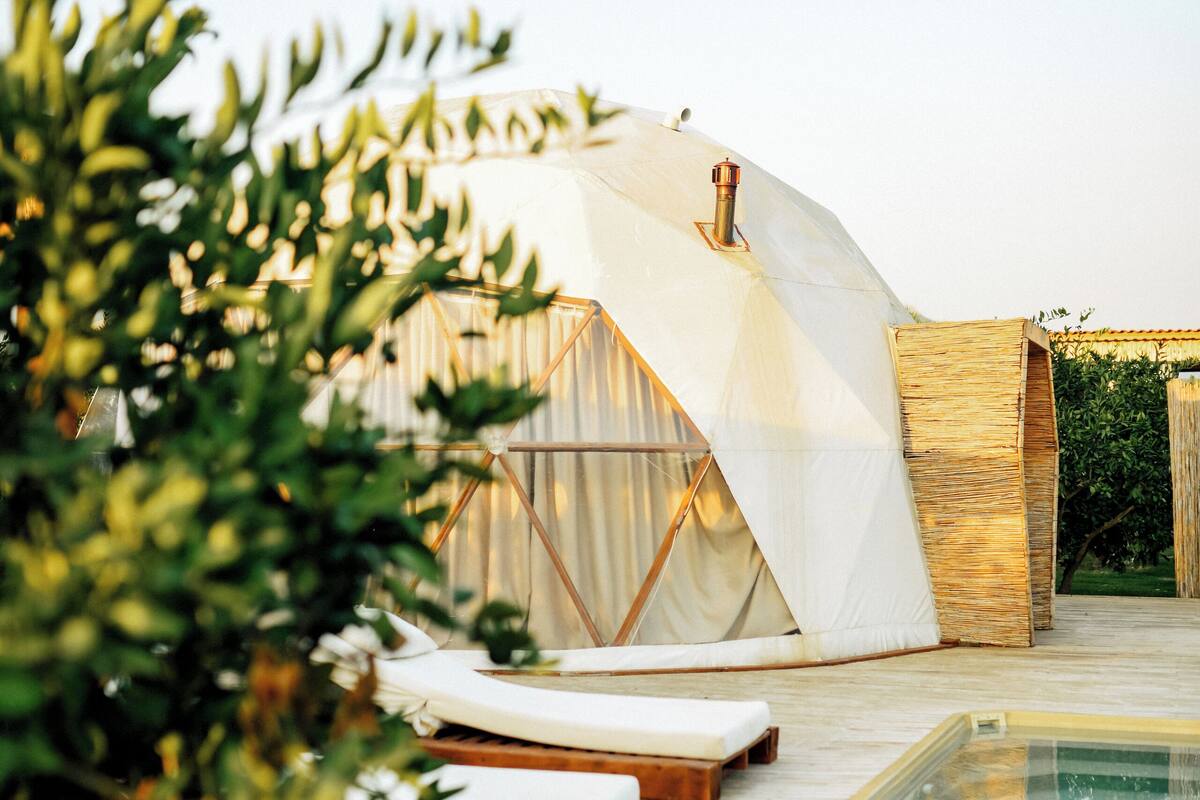 Une tente en forme de dôme dotée d’un jacuzzi en verre, entourée d’une terrasse en bois et de verdure.