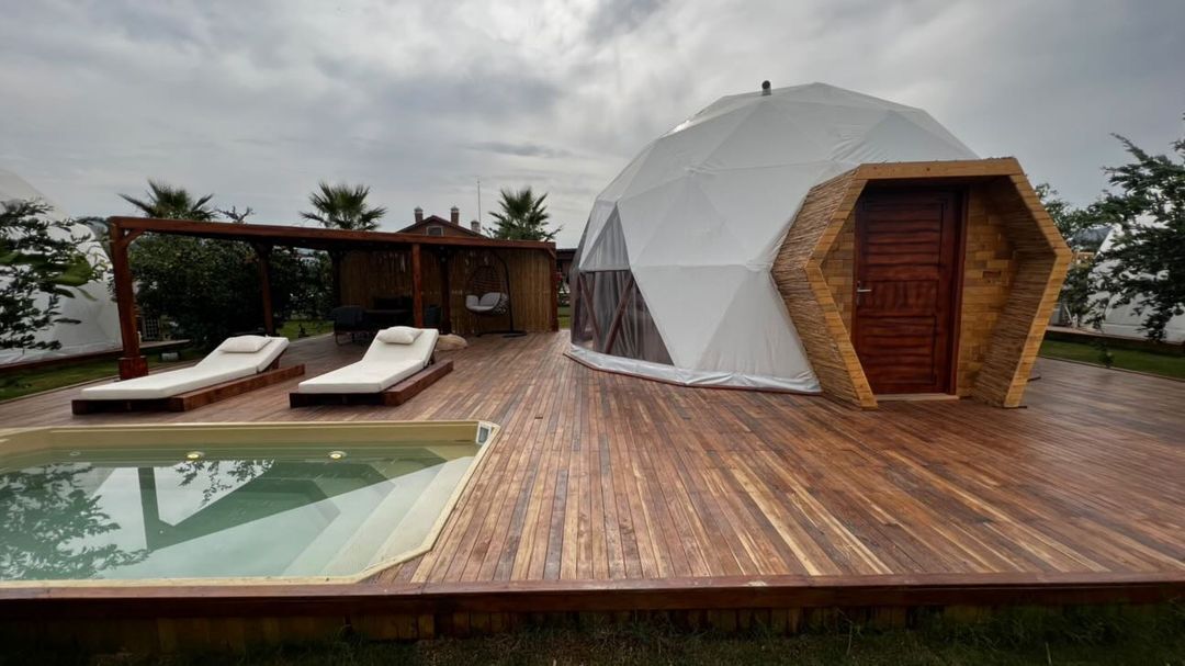 A dome-shaped tent with a wooden door, a pool, and lounge chairs on a wooden deck.