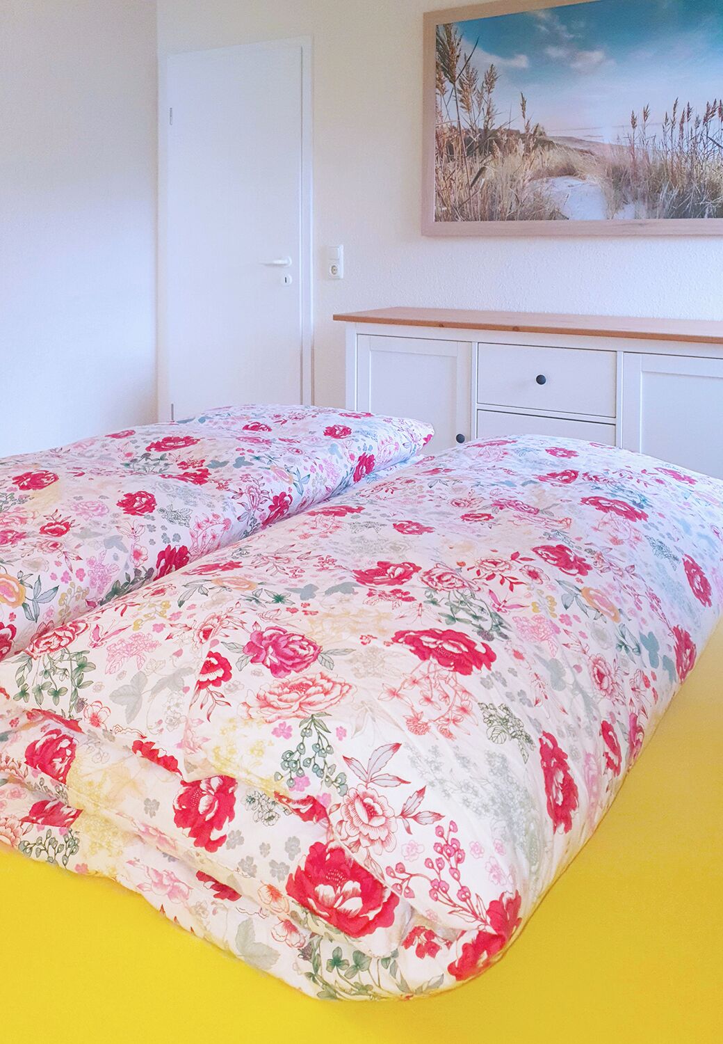 A bed with floral bedding, a white dresser, and a painting of a beach scene on the wall.