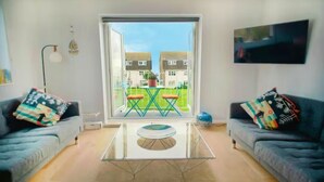 Ein modernes Wohnzimmer mit einem Glastisch, zwei Sofas mit bunten Kissen, einem großen Fenster mit Blick auf Häuser und einem Wand-Fernseher.