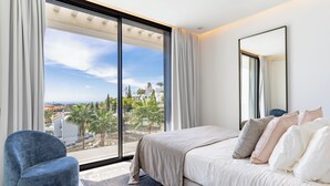 Ein modernes Schlafzimmer mit großem Fenster, einem Bett mit vielen Kissen, einem blauen Sessel und einem großen Spiegel.