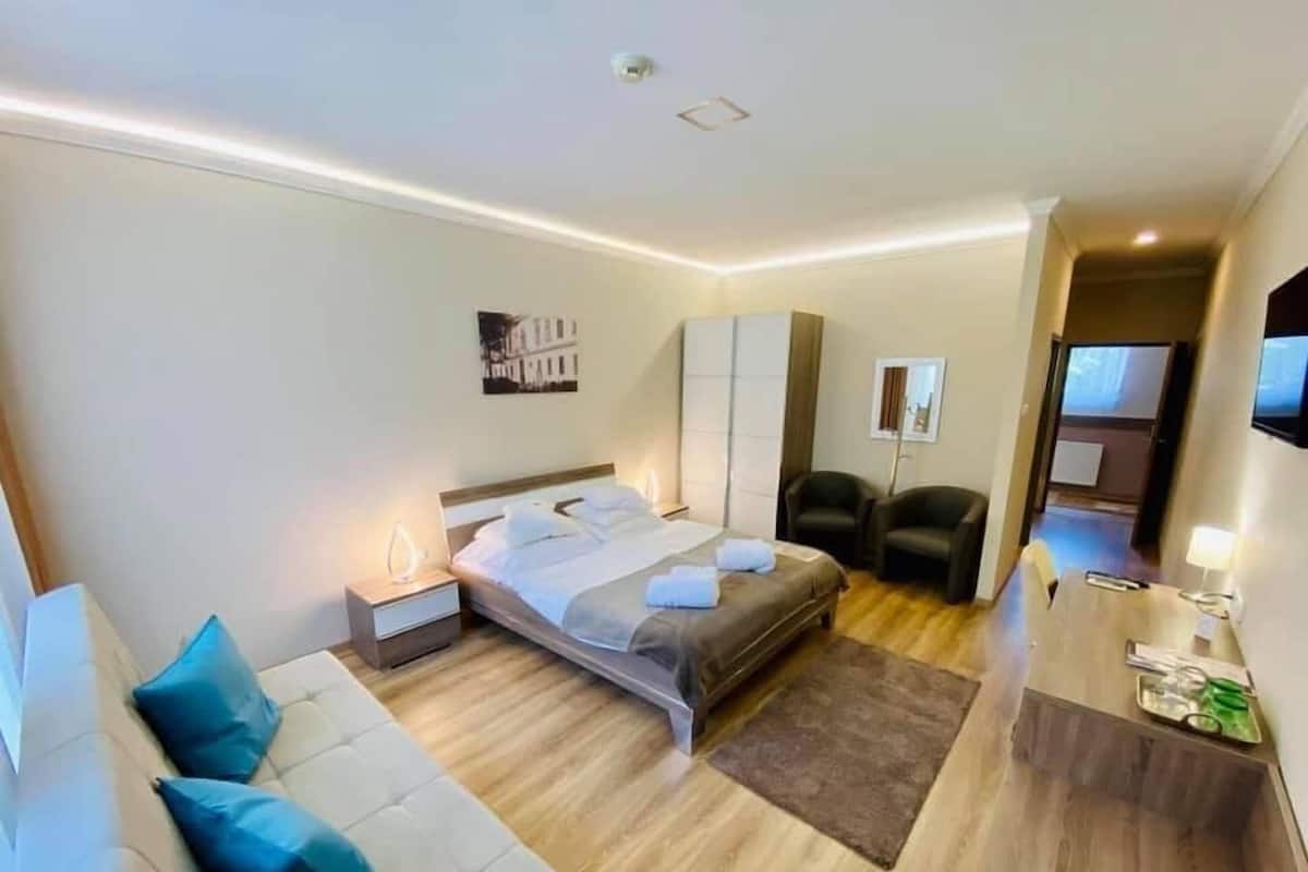 A hotel room with a bed, a sofa, a desk with a lamp, a chair, and a TV.