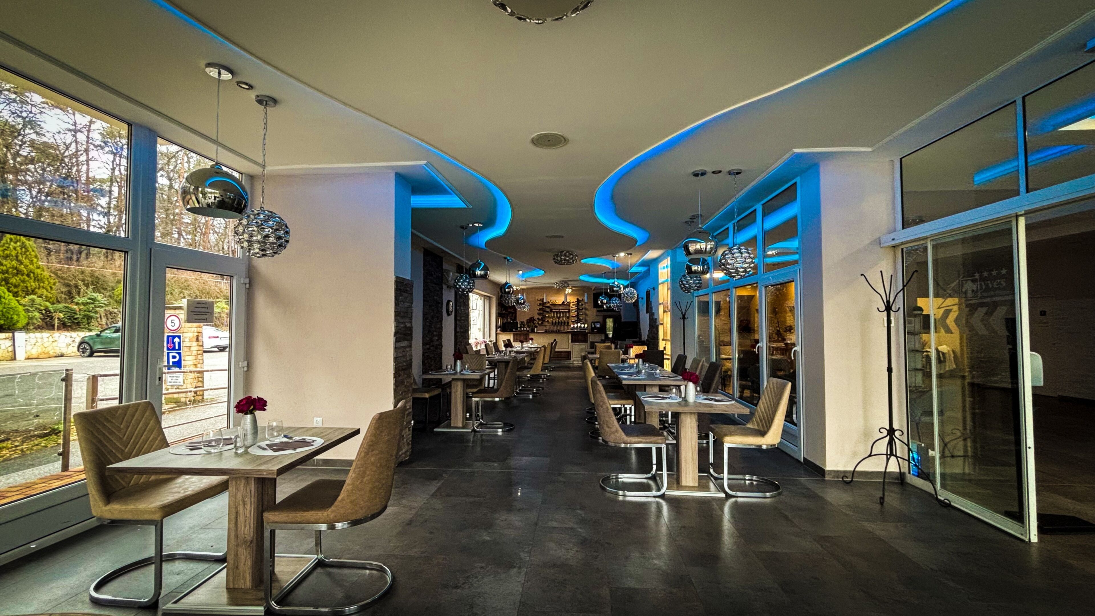 A modern restaurant interior with high ceilings, pendant lighting, and large windows offering views of the outdoors.