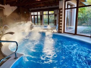 An indoor swimming pool with steam rising from the water, surrounded by large windows and a stone wall. - Fenyves Konferencia és Wellness Hotel (Baktalórántháza)