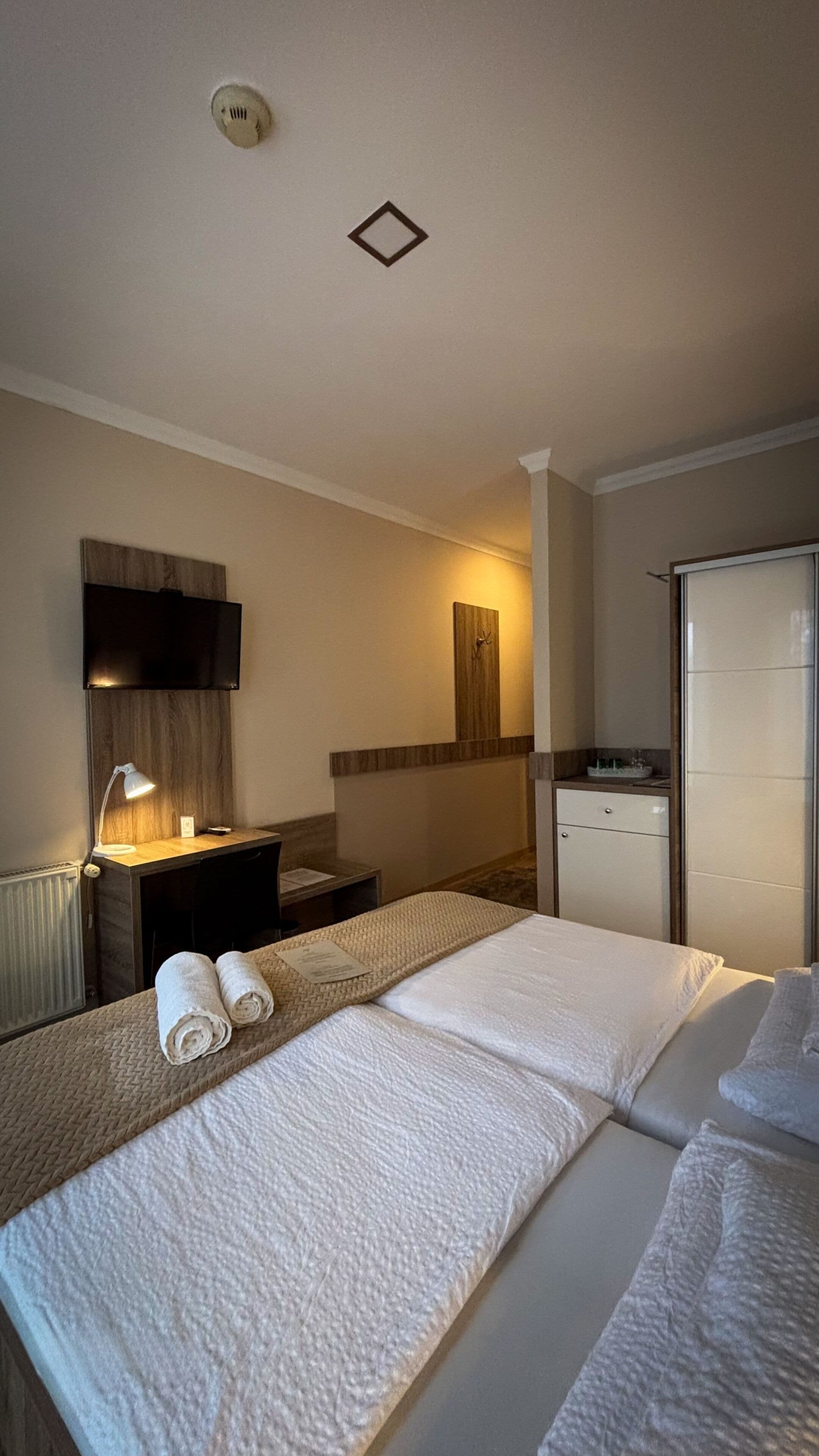 A hotel room with a bed, a desk with a lamp, a small table, a mirror, a wardrobe, and a wall-mounted TV.