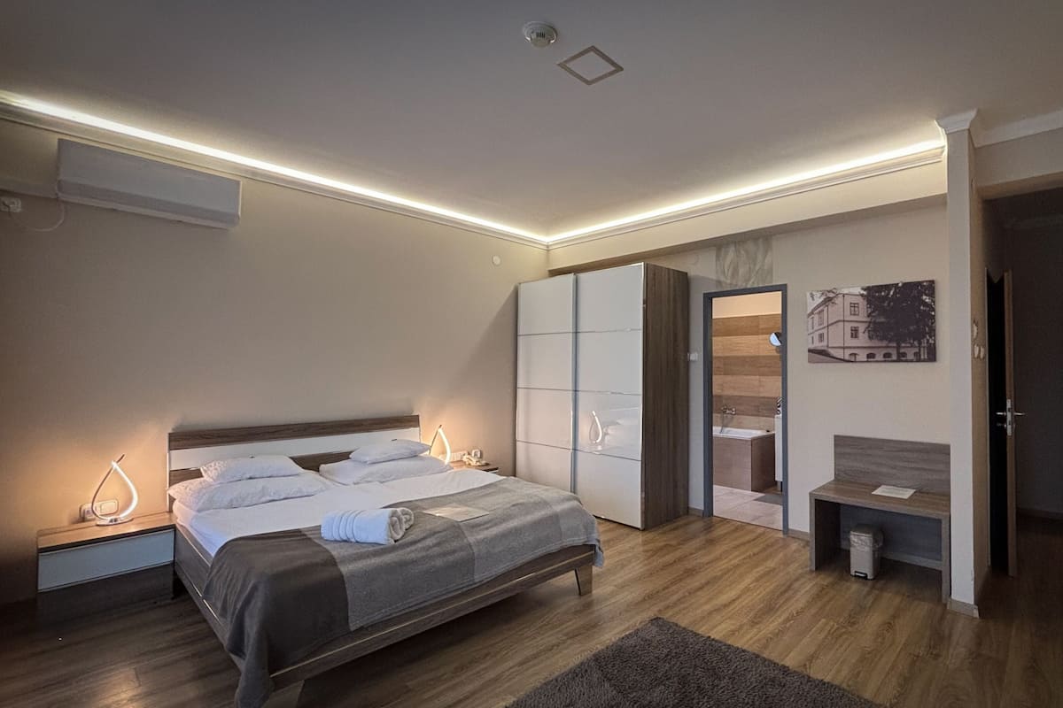 A modern bedroom with a bed, bedside lamps, a wardrobe, a bench, and a bathroom visible through an open door.