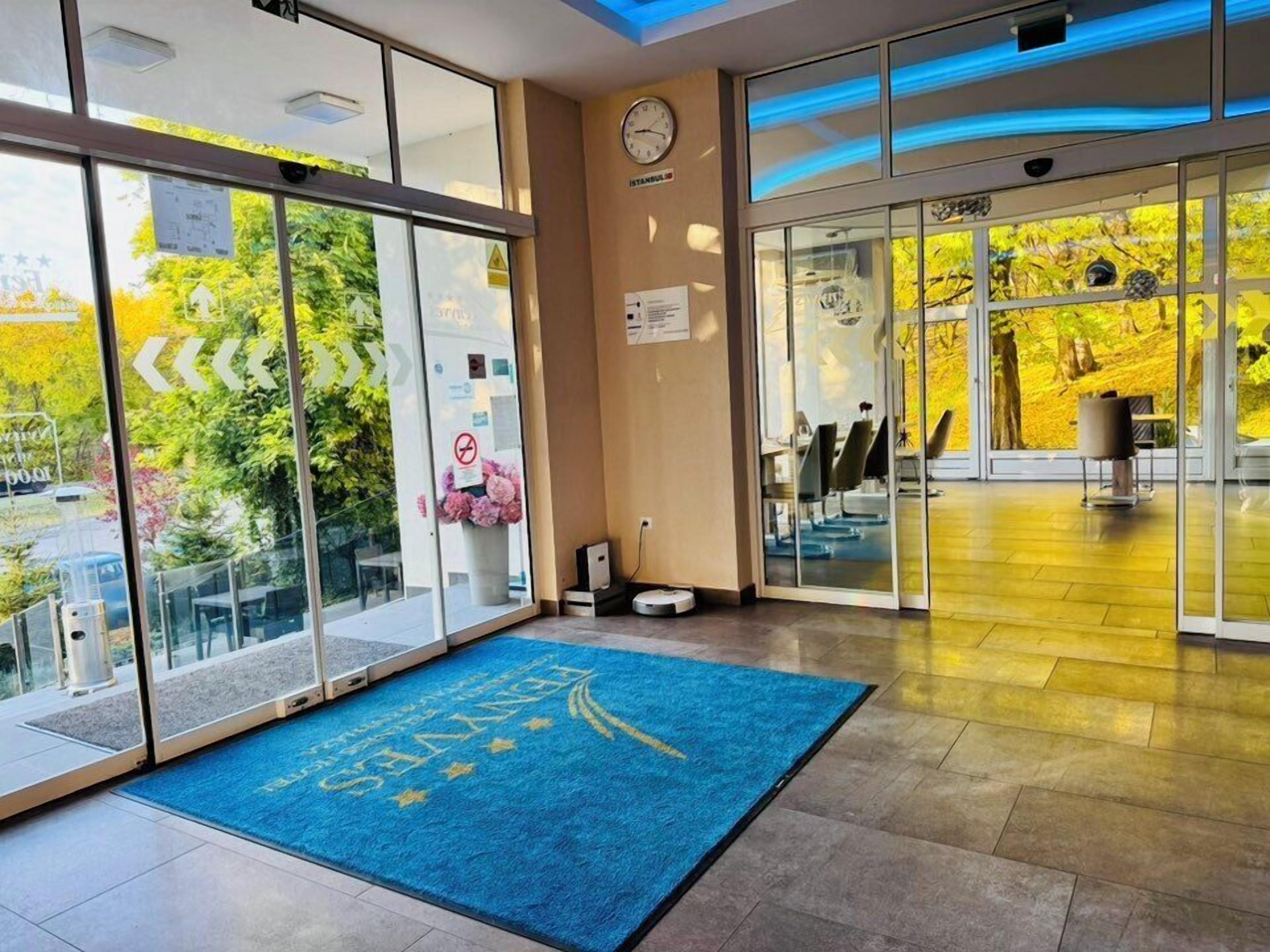 A hotel lobby with large glass doors, a blue carpet with text, a clock, and a view of trees.