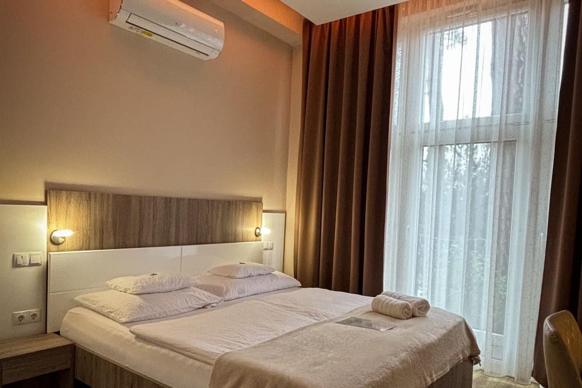 A hotel room with a bed, bedside tables, an air conditioner, a window with curtains, and a chair.
