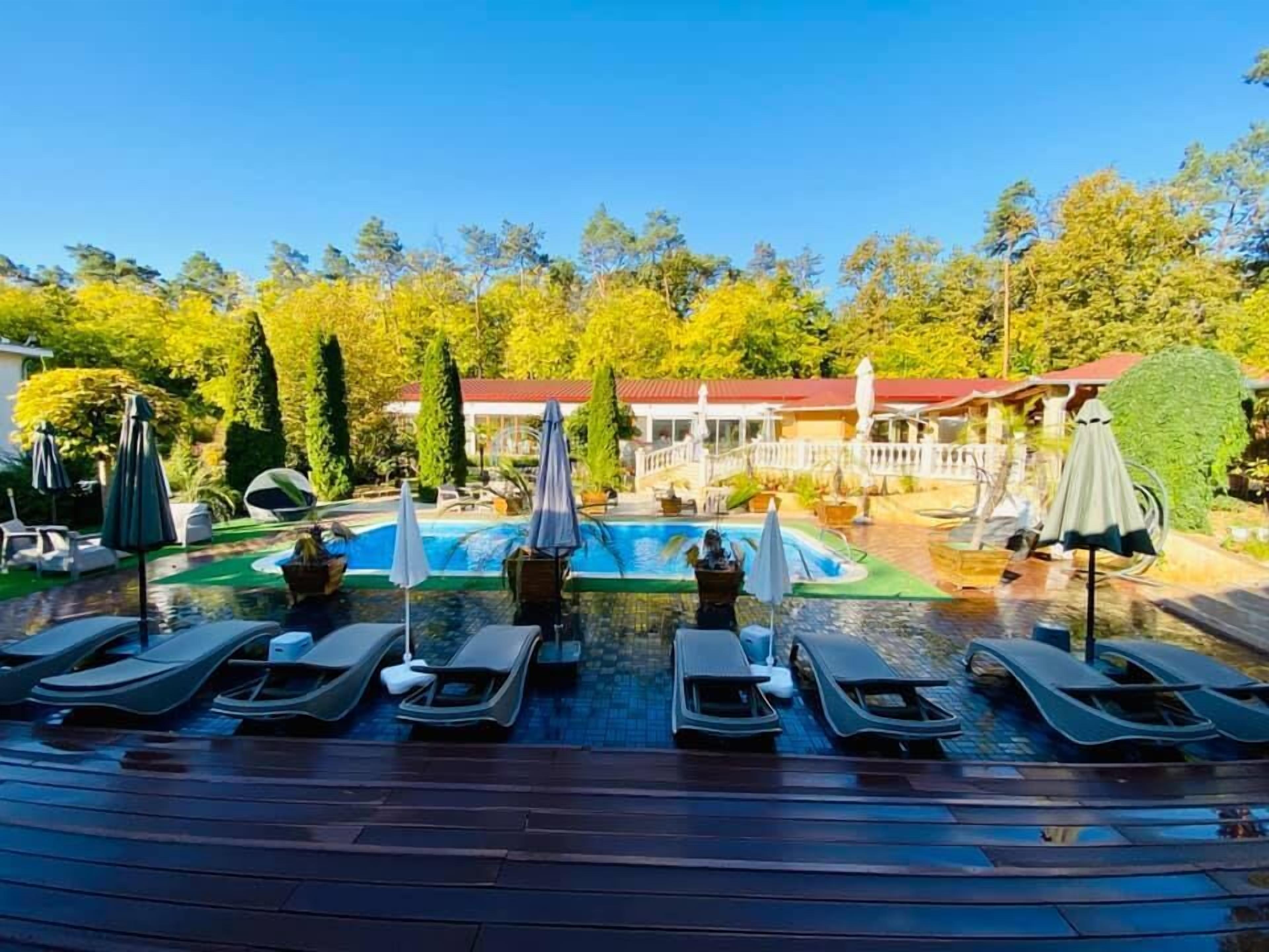 Una piscina con sillas de descanso y sombrillas, rodeada de árboles y un edificio de techo rojo.