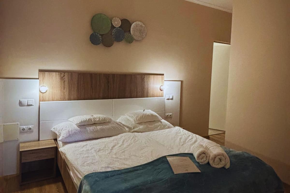 A bedroom with a wooden headboard, a bed with a blue cover, two bedside tables with lamps, and a wall-mounted light fixture.