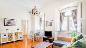 Ein Wohnzimmer mit Holzboden, einer Couch, einem Fernseher, einem Esstisch mit Stühlen und einer Kristallleuchter.