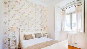 Ein Schlafzimmer mit blumigem Wandpapier, einem weißen Bett, zwei Nachttischen mit Lampen und einem Fenster mit Vorhängen.
