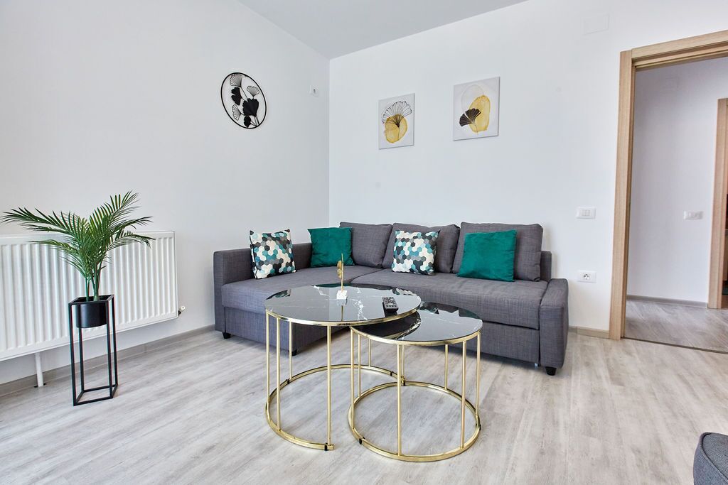 Un salon moderne avec un canapé gris, des coussins verts à motifs, une table basse en verre aux pieds dorés et une plante en pot.