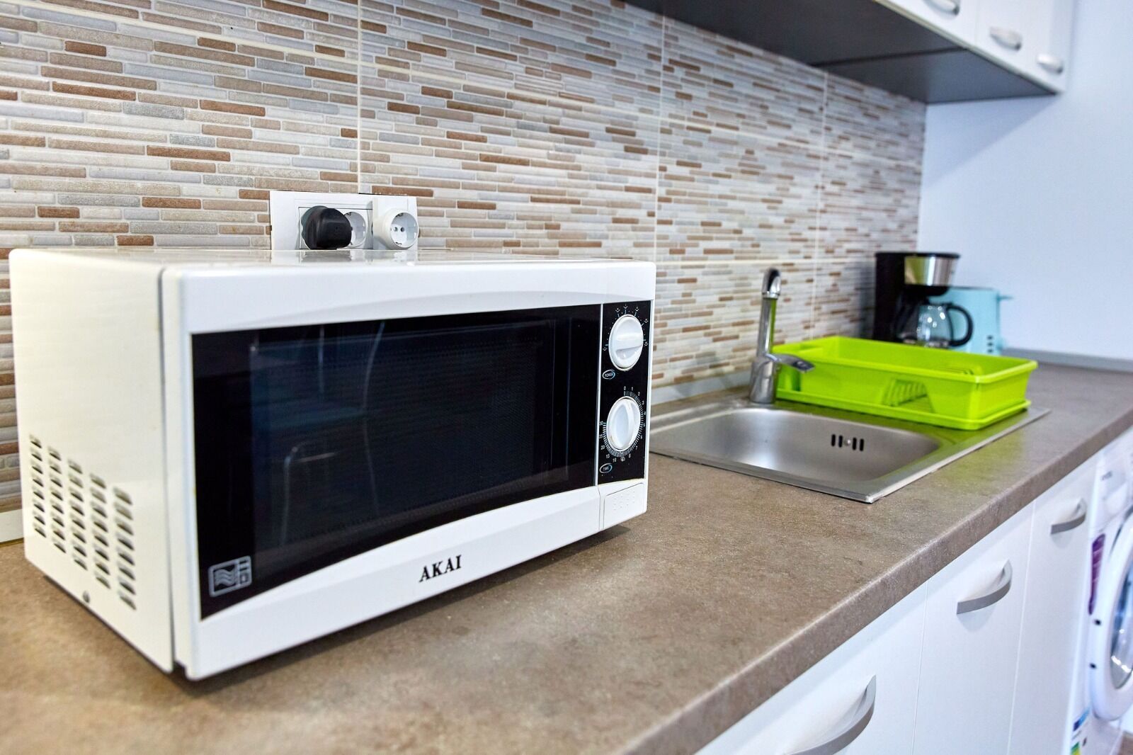 Un four à micro-ondes blanc, doté d’un écran numérique et de boutons de commande, est posé sur un plan de travail de cuisine, à côté d’un évier et d’un égouttoir à vaisselle vert.