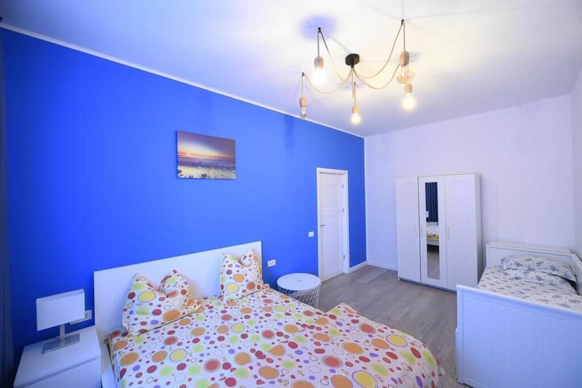 Quarto com parede azul, uma cama com colcha de retalhos coloridos, mesa de cabeceira branca e luminária no teto.