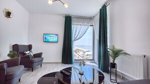 Un salón moderno con vistas al mar, una mesa redonda de cristal y un televisor de pantalla plana montado en la pared.