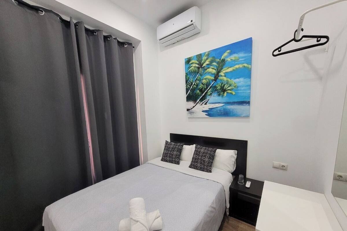 Un lit bien fait, recouvert d’une couverture grise et agrémenté de deux oreillers, une table de chevet blanche avec un téléphone, et un tableau représentant une scène de plage accroché au mur.