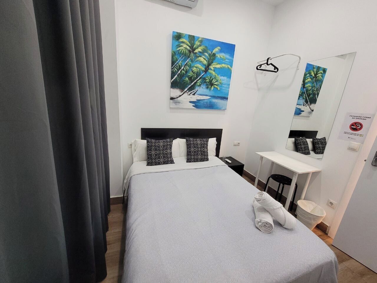 A hotel room with a bed, a desk, a chair, a mirror, and a painting of palm trees.