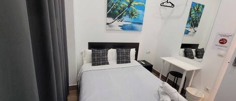 A hotel room with a bed, a desk, a chair, a mirror, and a painting of palm trees.