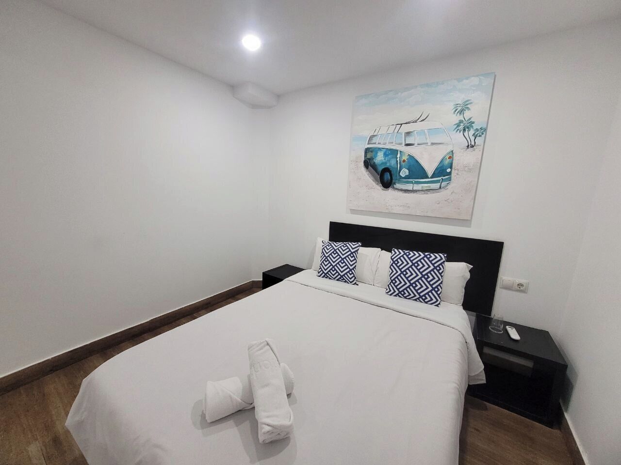Una cama bien hecha con sábanas y toalla blancos, una mesita de noche con un teléfono y un control remoto, y un cuadro de un coche en la playa sobre la cama.