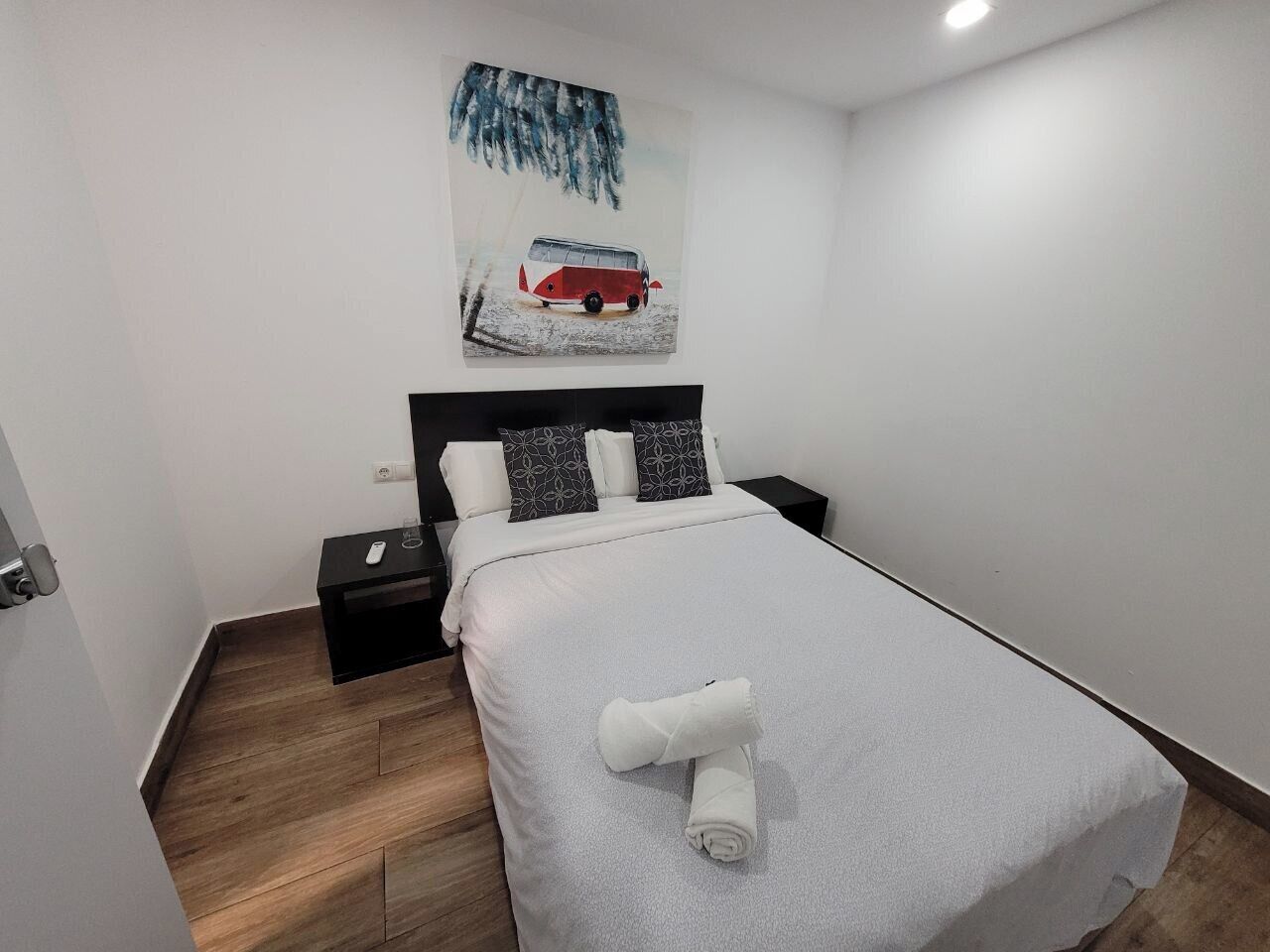 Una cama bien hecha con edredón blanco y una toalla enrollada, una mesita de noche con un teléfono y un cuadro colgado en la pared de un autobús rojo.