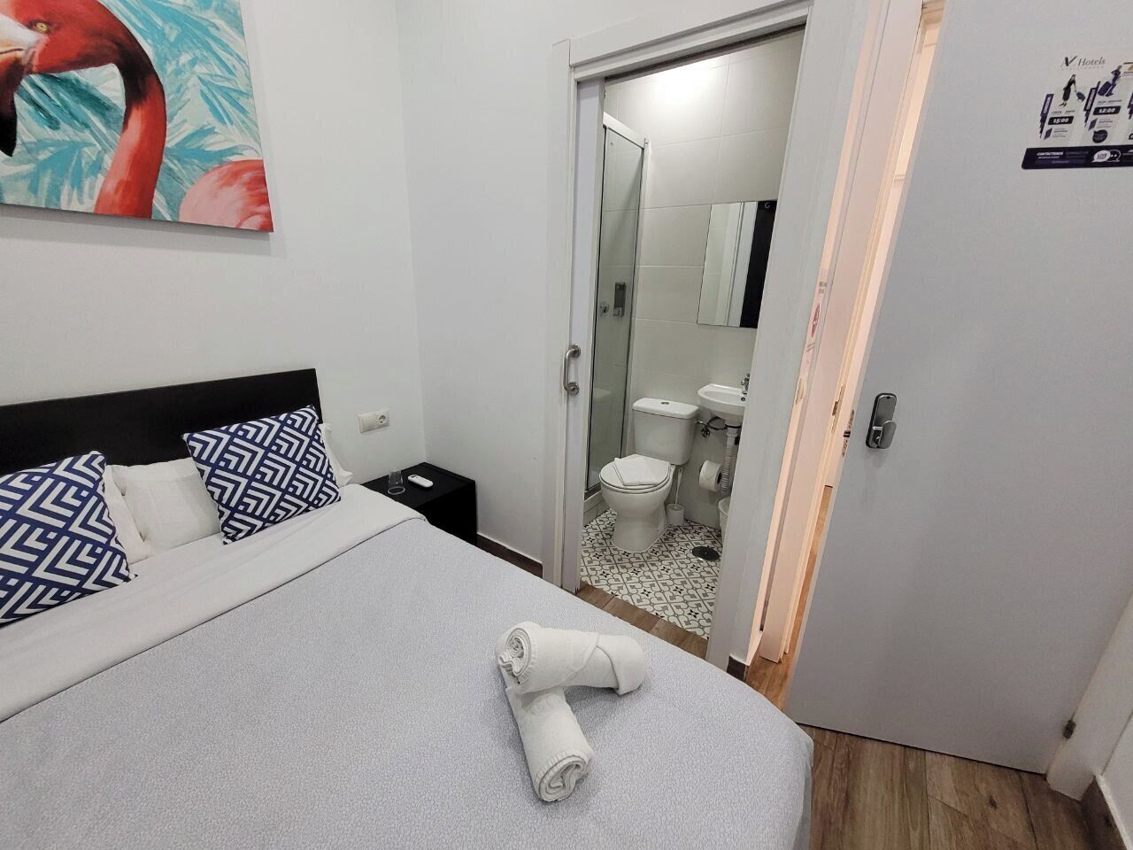 Una habitación de hotel con una cama, almohadones estampados, una toalla enrollada, una mesita de noche y un baño visible a través de una puerta abierta.
