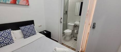 Una habitación de hotel con una cama, almohadones estampados, una toalla enrollada, una mesita de noche y un baño visible a través de una puerta abierta.