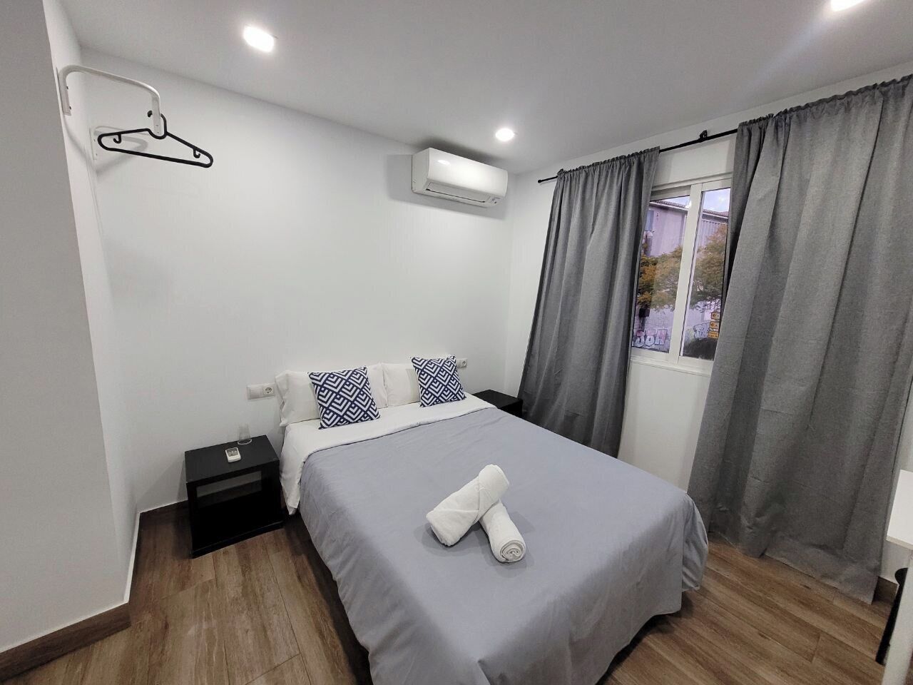 A hotel room with a bed, a nightstand, an air conditioning unit, a window with curtains, and a view of the outside.